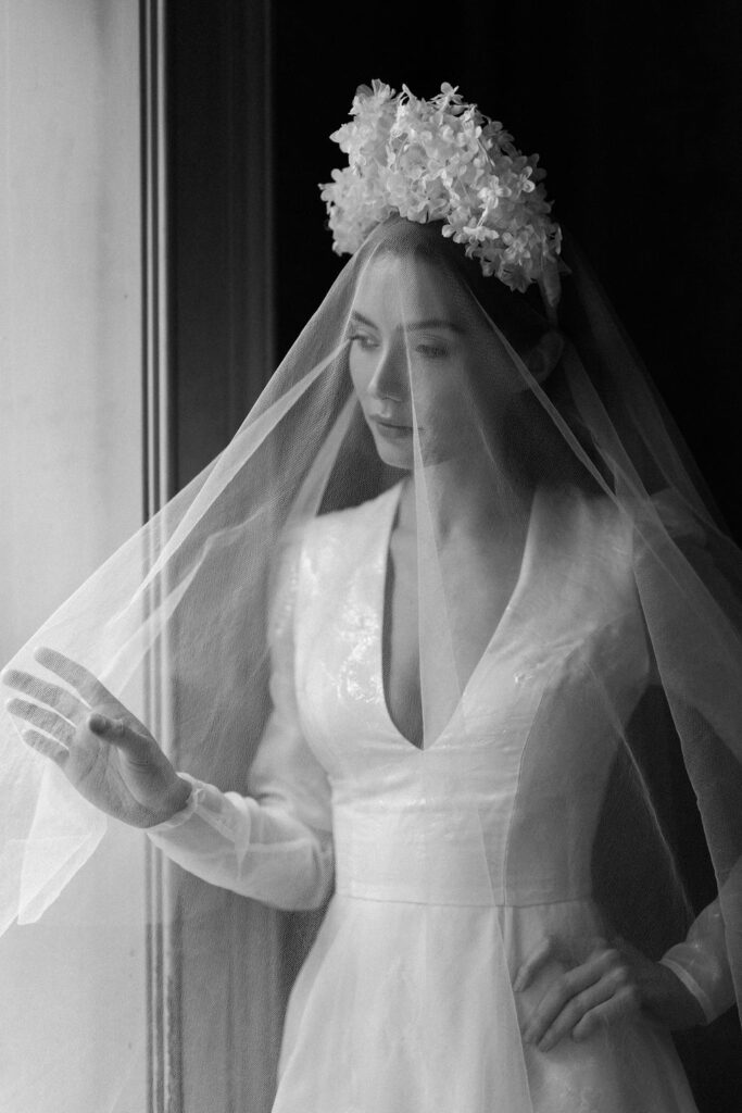 black and white portrait of bride lifting veil to look out window